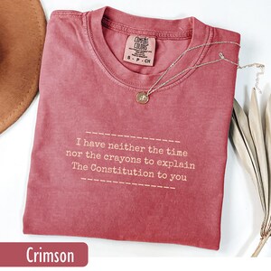 May include: Crimson-colored t-shirt with the text "I have neither the time nor the crayons to explain The Constitution to you." A silver necklace with a pendant rests on the shirt. The shirt is a size S.