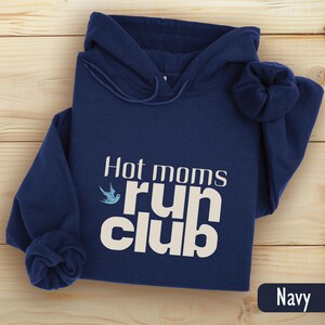 May include: A navy blue hoodie with the text "Hot moms run club" in white. A small blue bird graphic is to the left of the word "run". The word "Navy" is in a navy blue rectangle in the bottom right corner.