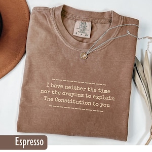 May include: Espresso-colored t-shirt with the text "I have neither the time nor the crayons to explain The Constitution to you." A silver necklace with a small pendant rests on the shirt. The shirt has a Comfort Colors tag.