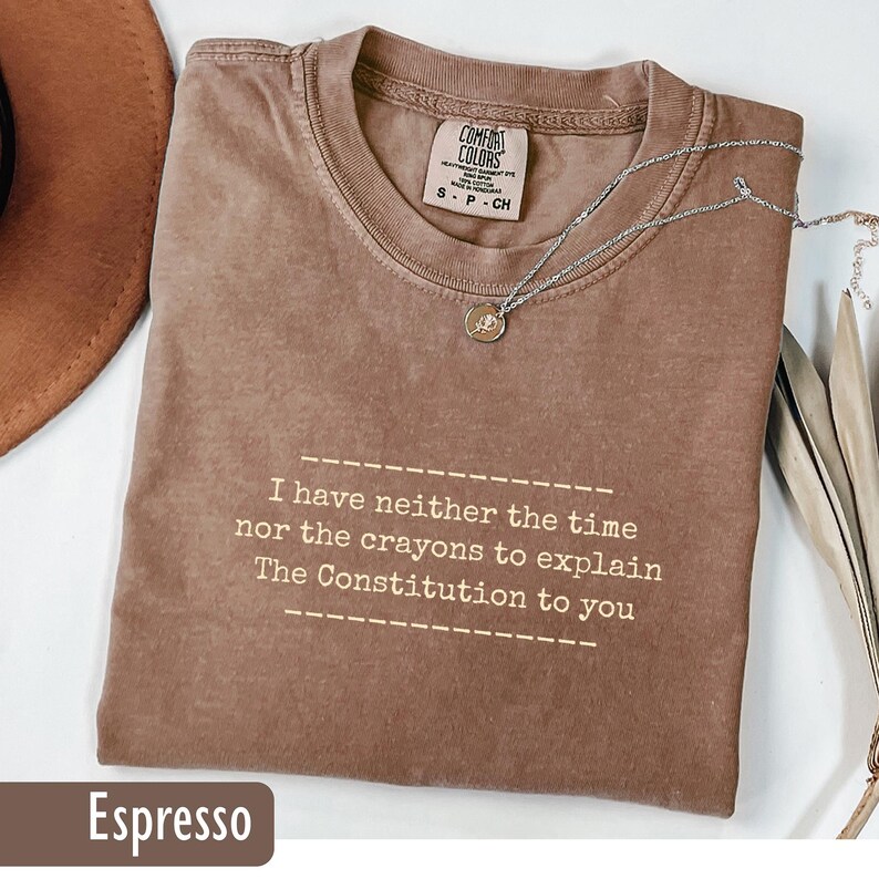 May include: Espresso-colored t-shirt with the text "I have neither the time nor the crayons to explain The Constitution to you." A silver necklace with a small pendant rests on the shirt. The shirt has a Comfort Colors tag.