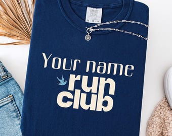 May include: A navy blue sweatshirt with the words "Your name run club" in white, and a small blue bird graphic. A silver necklace with a pendant is on the shirt. The words "True Navy" are at the bottom.