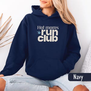 May include: Navy blue hooded sweatshirt with the text "Hot moms run club" in a cream color. The sweatshirt has a front pocket and a drawstring hood. The word "Navy" is displayed in a dark blue rectangle at the bottom right.