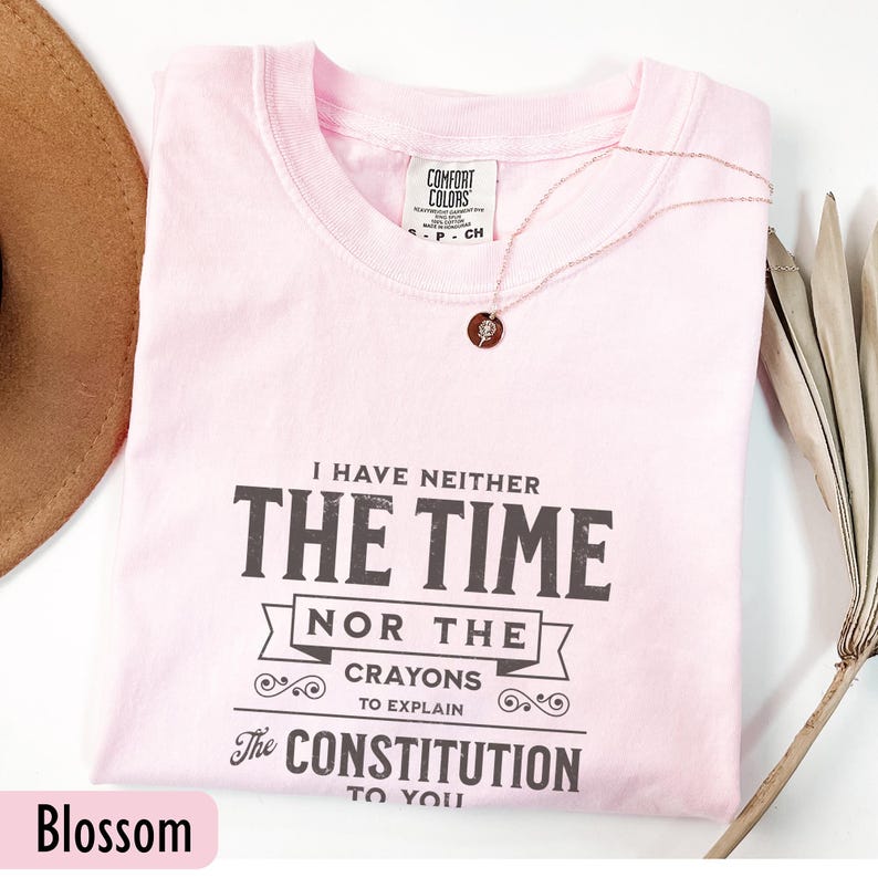May include: A light pink t-shirt with the text "I HAVE NEITHER THE TIME NOR THE CRAYONS TO EXPLAIN The CONSTITUTION TO YOU" in black. A rose gold necklace and a brown hat are also visible.