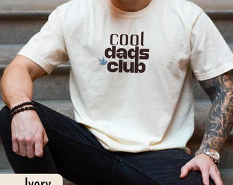 Könnte beinhalten: Elfenbeinfarbenes T-Shirt mit dem Text "cool dads club" in dunkelbrauner Schrift. Ein kleines blaues Vogel-Grafik befindet sich über dem Wort "club". Das Shirt ist aus weichem Material und hat eine lockere Passform.