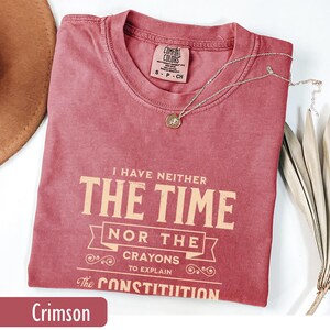 May include: Crimson-colored t-shirt with the text "I HAVE NEITHER THE TIME NOR THE CRAYONS TO EXPLAIN The CONSTITUTION." A silver necklace with a pendant is on the shirt. The shirt is a size small.