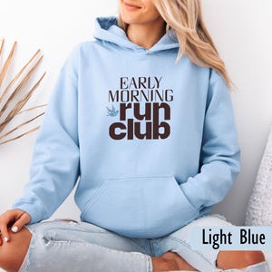 May include: Light blue hoodie with a front pocket and drawstring hood. The front of the hoodie features the text "EARLY MORNING RUN CLUB" in a brown font, with a small bird graphic. The bottom right corner of the image has the text "Light Blue".