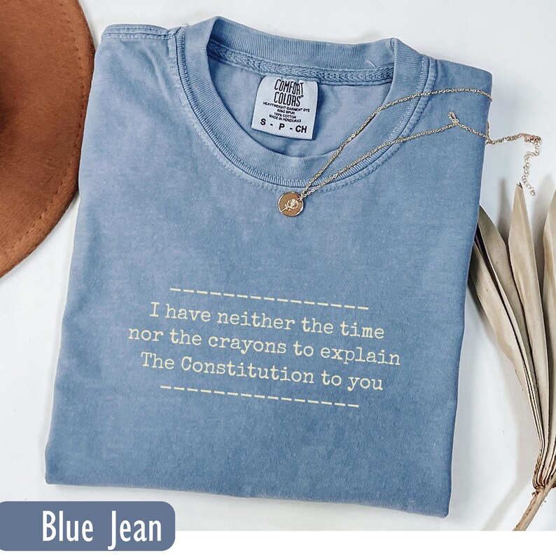 May include: A blue jean colored t-shirt with the text "I have neither the time nor the crayons to explain The Constitution to you." A gold necklace with a pendant rests on the shirt.