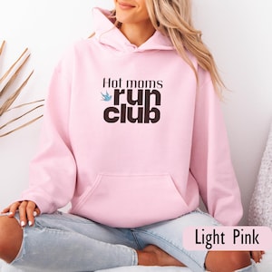 Puede incluir: Sudadera con capucha rosa claro con bolsillo delantero y capucha con cordón. La parte delantera presenta el texto "Hot moms run club" en negro y un gráfico de pájaro azul. Las palabras "Light Pink" están en la parte inferior.