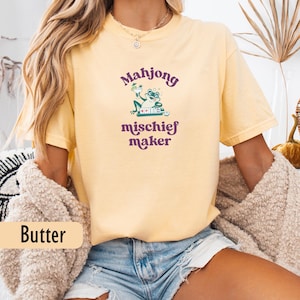 May include: Pale yellow t-shirt with a purple and green graphic. The graphic features the word "Mahjong" above a frog holding a drink, with the words "mischief maker" below. The shirt is a casual, comfortable style.