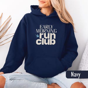 May include: Navy blue hooded sweatshirt with the text "EARLY MORNING run club" in white. The design includes a small blue bird graphic. The sweatshirt has a front pocket and a drawstring hood.