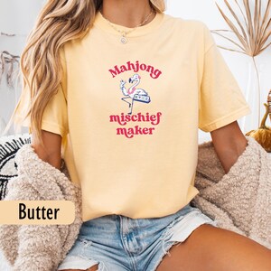 May include: Pale yellow t-shirt featuring a flamingo playing mahjong, with the words "Mahjong mischief maker" in pink. A relaxed fit, this tee is ideal for casual wear.