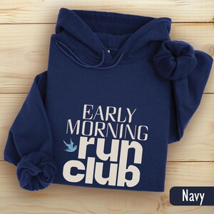 May include: Navy blue hooded sweatshirt with the text "EARLY MORNING run club" in white. A blue bird graphic is next to the words "run club". The sweatshirt is folded on a light wood surface.