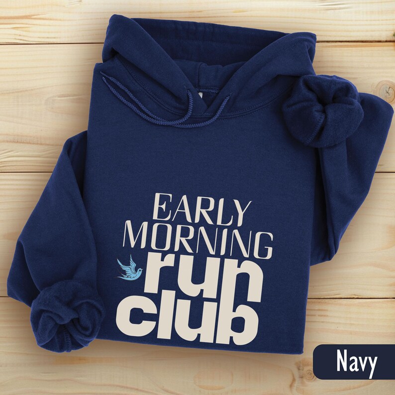 May include: Navy blue hooded sweatshirt with the text "EARLY MORNING run club" in white. A blue bird graphic is next to the words "run club". The sweatshirt is folded on a light wood surface.