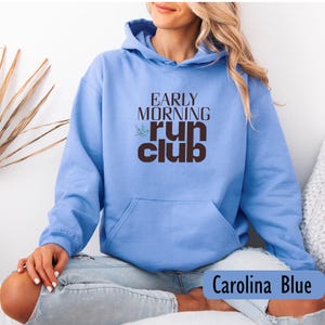 May include: Carolina blue hoodie with the text "EARLY MORNING RUN CLUB" in brown. The hoodie has a front pocket and a drawstring hood. The text is above a small green leaf design.