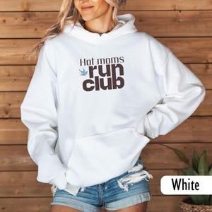 May include: White hooded sweatshirt with the text "Hot moms run club" in brown and blue. The sweatshirt has a front pocket and a drawstring hood. The text is in a casual font, and the design includes a small bird graphic.