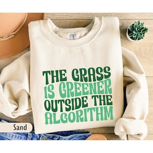 May include: A sand-coloured crewneck sweatshirt featuring the dark and light green text "THE GRASS IS GREENER OUTSIDE THE ALGORITHM" printed on the front. The garment has long sleeves and a ribbed collar, cuffs, and hem.