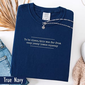 Könnte beinhalten: Ein marineblaues Sweatshirt mit dem weißen Text "To be clear, this was far from easy peasy lemon squeezy". Eine silberne Halskette mit Anhänger liegt auf dem Sweatshirt. Das Sweatshirt ist mit "True Navy" beschriftet.