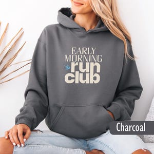 May include: Charcoal gray hoodie with a front pocket and drawstring hood. The hoodie features the text "EARLY MORNING RUN CLUB" in a cream-colored font, with a small blue bird graphic. The word "Charcoal" is displayed at the bottom right.