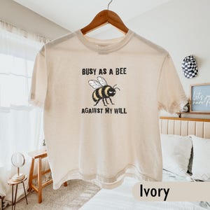 May include: An ivory short-sleeve t-shirt featuring a graphic of a bee with a grumpy expression. The text above the bee reads "BUSY AS A BEE" and the text below reads "AGAINST MY WILL" in a distressed black font. The shirt is hung on a wooden hanger.