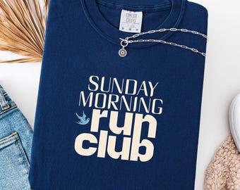 May include: A navy blue sweatshirt with the text "SUNDAY MORNING run club" in white. A small blue bird graphic is above the word "run". A silver necklace with a pendant is on the sweatshirt. The words "True Navy" are at the bottom.