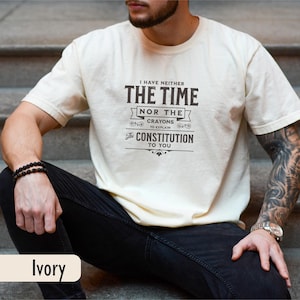 May include: Ivory-colored t-shirt with the text "I HAVE NEITHER THE TIME NOR THE CRAYONS TO EXPLAIN THE CONSTITUTION TO YOU" in black. The person is wearing black jeans and a black beaded bracelet.