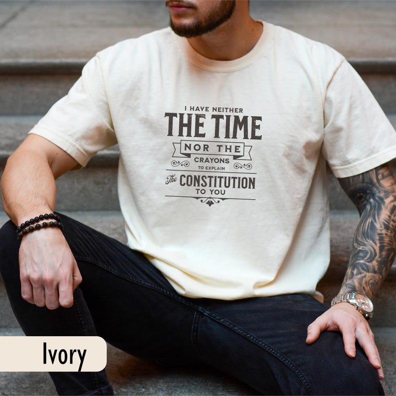 May include: Ivory-colored t-shirt with the text "I HAVE NEITHER THE TIME NOR THE CRAYONS TO EXPLAIN THE CONSTITUTION TO YOU" in black. The person is wearing black jeans and a black beaded bracelet.