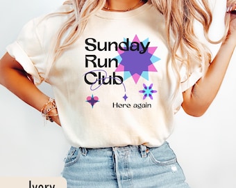 May include: Ivory-colored t-shirt with the text "Sunday Run Club" in black, with a purple and blue starburst design. The words "Here again" are below the graphic. The shirt is paired with blue jeans.