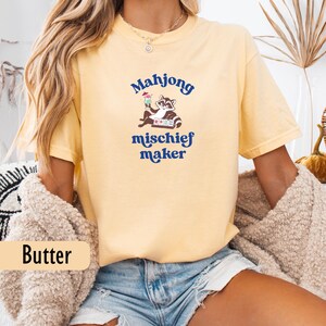 May include: Pale yellow t-shirt with the text "Mahjong mischief maker" in blue. The graphic shows a raccoon with a cocktail and a mahjong tile. The shirt has a loose fit.
