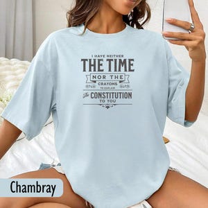 May include: Light blue chambray t-shirt with the text "I HAVE NEITHER THE TIME NOR THE CRAYONS TO EXPLAIN The CONSTITUTION TO YOU" in dark gray. The shirt has short sleeves and a relaxed fit.