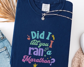 May include: A navy blue sweatshirt with the text "Did I tell you I ran a Marathon?" in colorful, playful lettering. The shirt has a crew neck and a Comfort Colors tag. A silver necklace with a small pendant is draped across the shirt.
