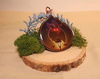 May include: A teardrop-shaped keychain featuring a cartoon moth with a glowing red face and a lamppost. The keychain has a red and brown border and a silver keyring. The keychain is displayed on a wooden slice with green moss and blue decorative sprigs.