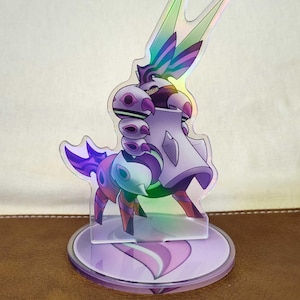 Op de afbeelding: Een holografische acryl standee met een gestileerd wezen in paarse en teal tinten. Het wezen heeft een uniek ontwerp met meerdere oogvormige vormen en een cape. De standee staat op een cirkelvormige basis.