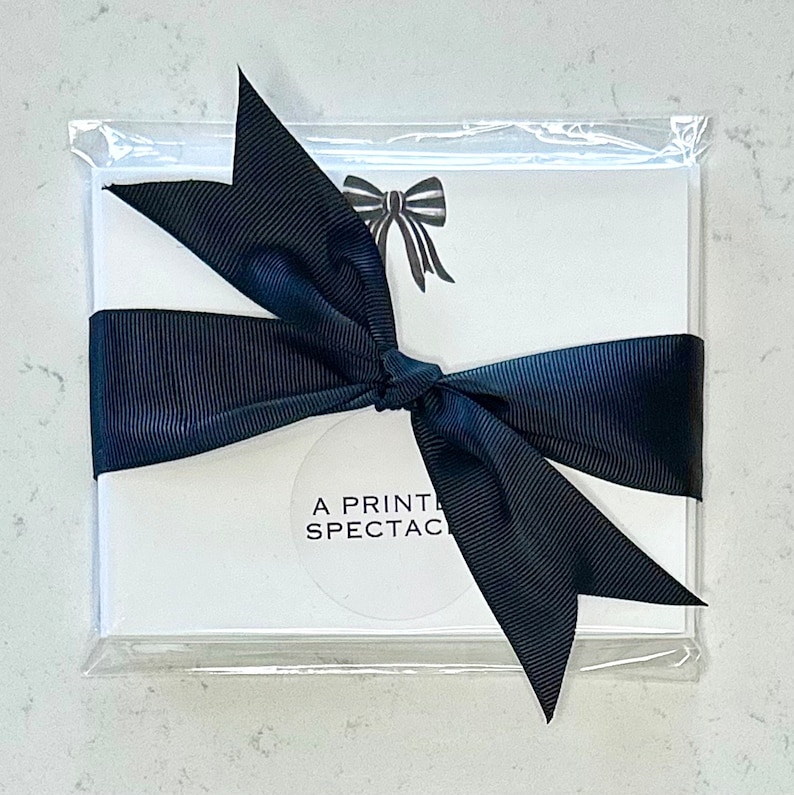 Peut inclure: Un ensemble de cartes blanches avec un n&oelig;ud noir et le texte "A PRINT SPECTACLE" sur le devant, envelopp&eacute; d'un ruban noir et emball&eacute; dans un sac en plastique transparent.