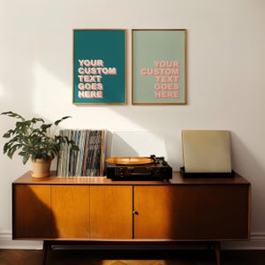 Op de afbeelding: Een houten kast in mid-century modern stijl met een platenspeler, vinylplaten en twee ingelijste prints. De prints hebben gouden frames en de tekst "YOUR CUSTOM TEXT GOES HERE" in roze. Een potplant staat op de kast.