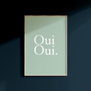 May include: Framed art print with a light green background and the words "Oui Oui" in white, sans-serif font. The frame is a thin, gold color. The artwork is displayed against a dark blue wall.