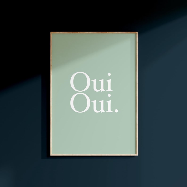 May include: Framed art print with a light green background and the words "Oui Oui" in white, sans-serif font. The frame is a thin, gold color. The artwork is displayed against a dark blue wall.