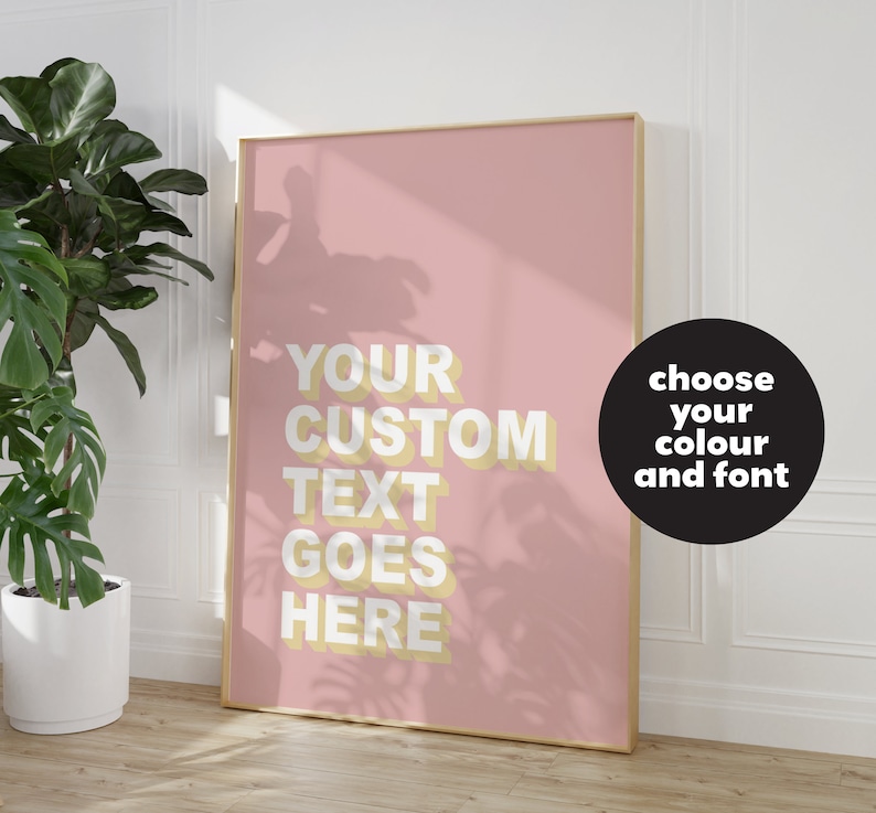May include: A pink poster with a gold frame and the text "YOUR CUSTOM TEXT GOES HERE" in yellow. The poster is on a white wall with a green plant in a white pot in the foreground. A black circle with white text says "choose your colour and font".