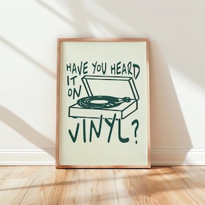 Könnte beinhalten: Ein gerahmter Druck mit einer grünen Illustration eines Plattenspielers und dem Text "HAVE YOU HEARD IT ON VINYL?". Das Kunstwerk befindet sich auf einem hellgrünen Hintergrund, eingerahmt in Naturholz, vor einer weißen Wand.