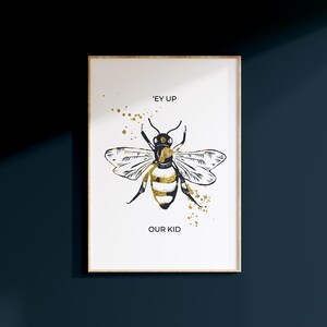 Puede incluir: Impresi&oacute;n art&iacute;stica enmarcada con una ilustraci&oacute;n de abeja en negro y dorado. El texto dice "'EY UP OUR KID". Salpicaduras de pintura dorada acent&uacute;an el dise&ntilde;o. El marco es de color madera clara.