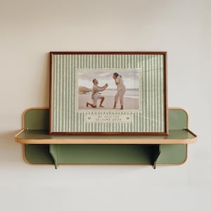 Peut inclure: Une impression rectangulaire encadrée avec un fond vert sauge et des rayures verticales blanches. Le centre présente une photographie d'une demande en mariage sur une plage, avec le texte "WE SAID YES 12 JUNE 2026" sous l'image.