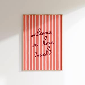 May include: A framed print with a coral and white striped background. The text "welcome, we have snacks" is written in a dark red script. The frame is a light wood color.