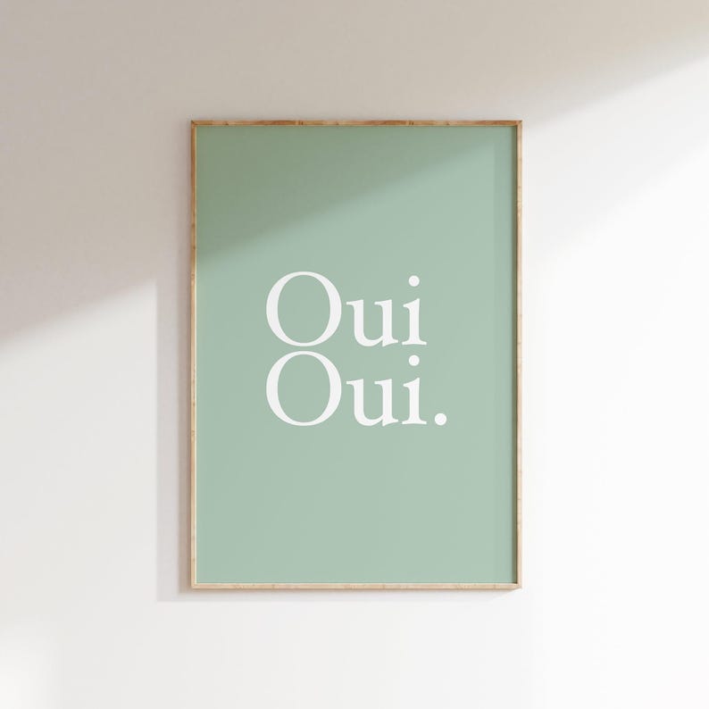 May include: A framed art print featuring the words "Oui Oui" in white, centered on a soft green background. The frame is a light wood color. The print is displayed on a white wall.