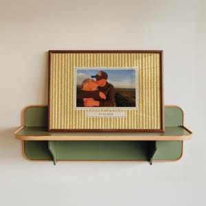 Può includere: Una cornice rettangolare con bordo in legno scuro contenente una fotografia di due persone che si abbracciano all'aperto al tramonto. La cornice presenta uno sfondo giallo senape con strisce verticali bianche e la data 01.04.2026 stampata sotto.