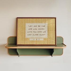 Op de afbeelding: Een rechthoekige ingelijste print met een mosterdgele achtergrond en verticale witte strepen. In het midden staat de tekst "LET ME BE THE ONE WHO SHINES WITH YOU AND WE CAN SLIDE AWAY" binnen een gestippelde rand, met muziekspeler-iconen eronder.