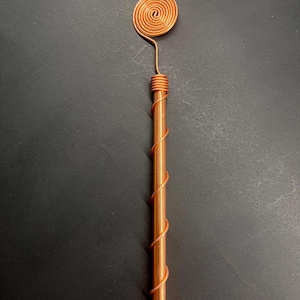 May include: A copper wand with a spiral design. The wand features a cylindrical body wrapped with copper wire, and a spiral at the top. The wand is made of copper and has a shiny finish.