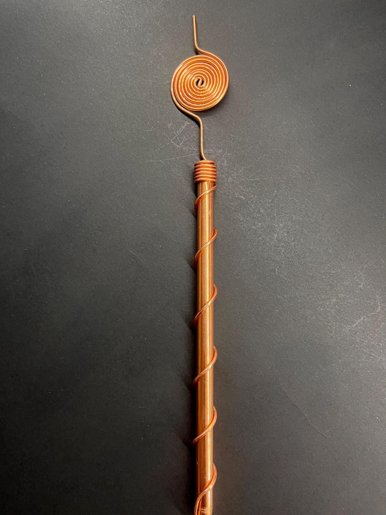 May include: A copper wand with a spiral design. The wand features a cylindrical body wrapped with copper wire, and a spiral at the top. The wand is made of copper and has a shiny finish.