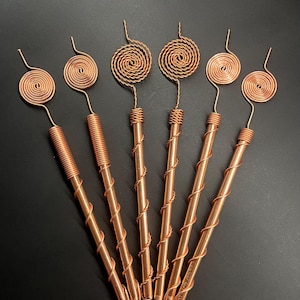 May include: Copper wire art pieces with spiral and coil designs. The pieces have a copper finish and are arranged on a dark surface. The tops feature a spiral design, while the stems have a wrapped or coiled appearance.