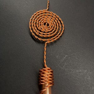 May include: A copper-colored, handmade pendant with a spiral design. The pendant features a coiled spiral at the top, connected to a wrapped cylindrical base. The metal has a warm, reddish-brown hue.