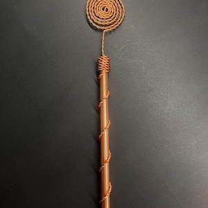 May include: A copper-colored wand with a spiral design at the top and a wrapped handle. The wand's top features a circular spiral, and the handle is wrapped with copper wire. The wand is a unique, handcrafted item.
