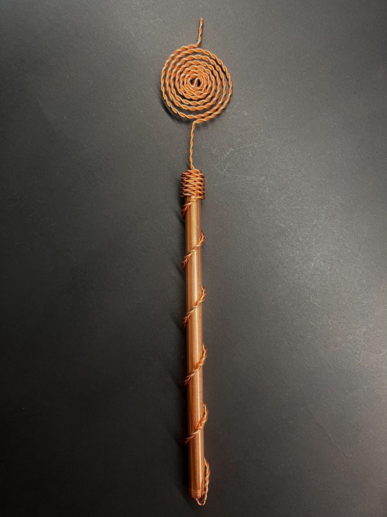 May include: A copper-colored wand with a spiral design at the top and a wrapped handle. The wand's top features a circular spiral, and the handle is wrapped with copper wire. The wand is a unique, handcrafted item.